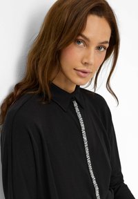 Frau mit langen braunen Haaren trägt ein schwarzes Hemd mit Kragen, das an der Vorderseite eine vertikale Reihe kleiner weißer Perlen oder Strasssteine aufweist.