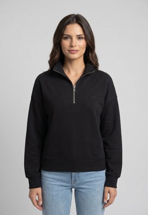 Mujer con cabello castaño que lleva un jersey corto negro con medio cierre y vaqueros azul claro, de pie frente a un fondo gris liso.