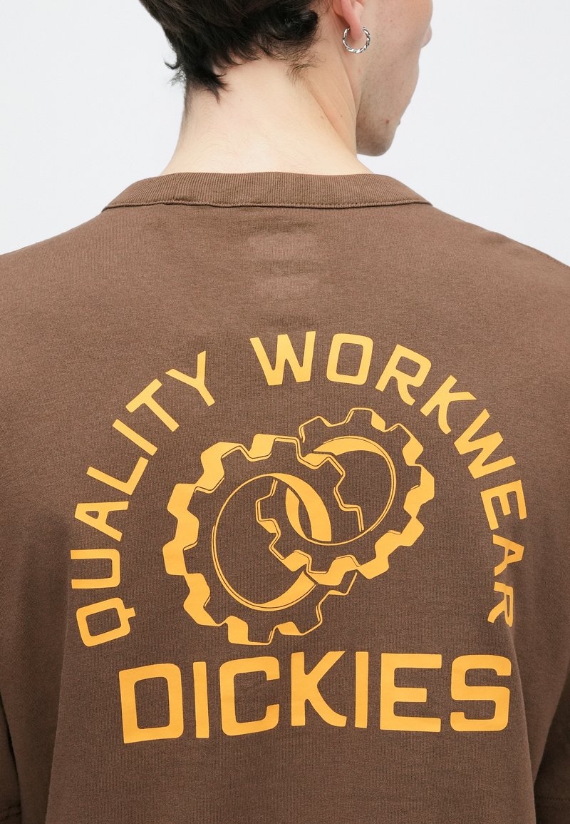 Personne portant une chemise marron avec le texte jaune « Quality Workwear Dickies » et des graphiques de rouages superposés au dos.