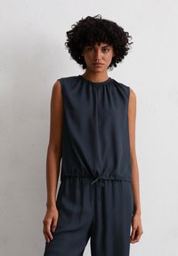 Frau mit lockigem Haar, die ein ärmelloses dunkelblaues Oberteil und eine passende Hose trägt, steht vor einer schlichten weißen Wand.