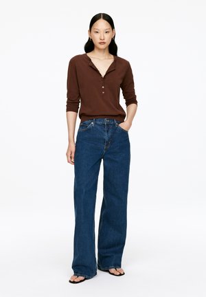 Brown henley shirt with three buttons, paired with high-waisted, loose-fit blue jeans. Model stands with hands in pockets, wearing black sandals.