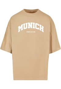 Beež üle suuruse T-särk, mis on valmistatud puuvillast, sisaldades valget kumerat teksti, mis ütleb "MUNICH MERCH•DE" rinna piirkonnas.
