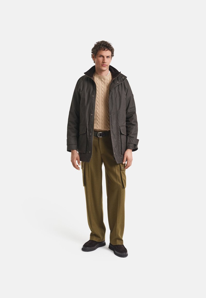 Dunkelbraune wasserdichte Jacke mit Kapuze, getragen über einem cremefarbenen Kabelstrickpullover und olivgrünen Cargo-Shorts. Dunkle Schuhe runden das Outfit ab.