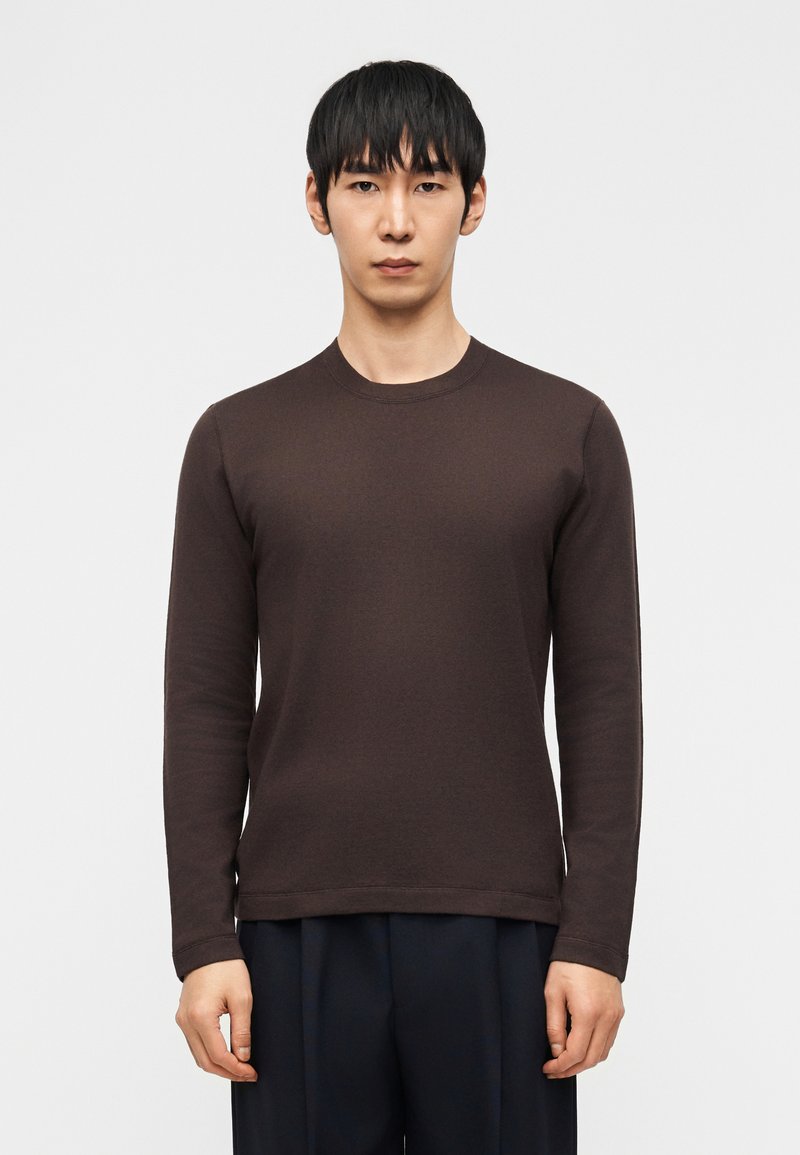 Άνδρας με ίσια μαύρα μαλλιά, φορώντας σκούρο καφέ μακρυμάνικη μπλούζα με στρογγυλή λαιμόκοψη και σκούρο παντελόνι, στέκεται μπροστά σε λευκό απλό φόντο.