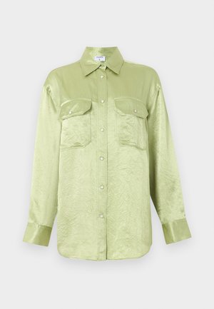 Žena nosi svetlozelenú oversized saténovú košeľu, čierne široké nohavice, čierne špicaté baleríny a drží čiernu kabelku.