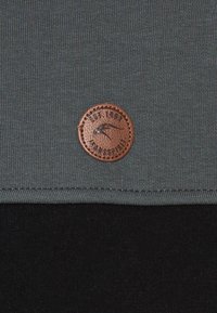 Tessuto grigio con superficie testurizzata, dotato di una patch rotonda in pelle marrone con un design e testo impresso, cucita sul bordo inferiore.