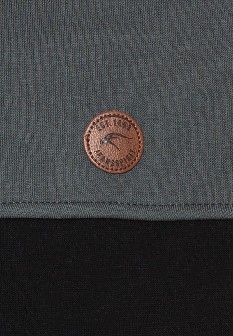 Tissu gris avec une surface texturée, comportant un patch en cuir marron rond avec un design en relief et du texte, cousu sur le bord inférieur.