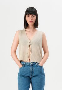 Vrouw met steil zwart haar, gekleed in een beige mouwloze gebreide vest, geknoopt vooraan, en blauwe high-waisted jeans met haar handen in haar zakken.