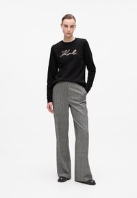 Femme debout avec une main dans la poche, portant un sweatshirt noir avec le texte "Karl" et un pantalon large gris avec des chaussures noires.
