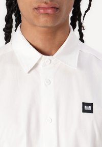 Hvit bomulls skjorte med knapper som har et svart merket med "WEEKEND OFFENDER"-logo. Klassisk krage, enkel tekstur, og fem synlige knapper.