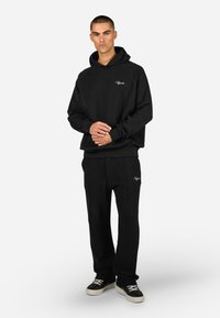 Μαύρη φόρμα με κουκούλα και μπροστινή τσέπη, διακοσμημένη με κεντημένο λογότυπο, συνδυασμένη με υφασμάτινες μαύρες φόρμες. Τα αθλητικά παπούτσια ολοκληρώνουν την καθημερινή ενδυμασία.