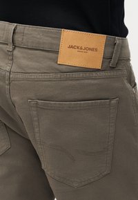 Καφέ παντελόνι τζιν που δείχνει την πίσω τσέπη και ετικέτα δερμάτινου λογότυπου σε ταμπά χρώμα με την επωνυμία "JACK & JONES", συνδυασμένο με μαύρο πουκάμισο.