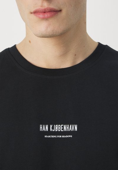 Tricou din bumbac negru cu guler rotund. Partea din față prezintă text alb: "HAN KJØBENHAVN" și "CĂUTÂND UMBRIRI". Textură moale.