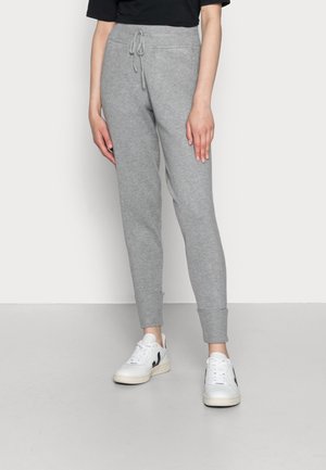 Osoba nosząca szare spodnie dresowe z wiązanym pasem i prążkowanymi mankietami, w połączeniu z białymi sneakersami z czarnymi akcentami, stojąca na szarej podłodze.