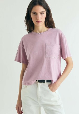 Mujer con cabello largo y oscuro que lleva una camisa de manga corta color púrpura claro con tachuelas, metida dentro de un pantalón blanco de cintura alta con un cinturón negro.