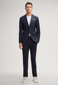 Abito navy con due bottoni, revers a punta e due tasche frontali, abbinato a una maglietta bianca e sneaker bianchi. Tessuto a trama liscia.