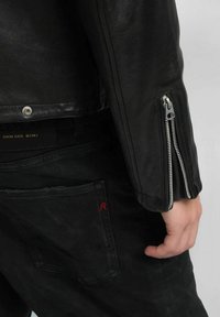 Zwarte leren jas met zilveren ritsen op de mouwen, gecombineerd met zwarte broek met een rood accentstiksel op de achterzak.