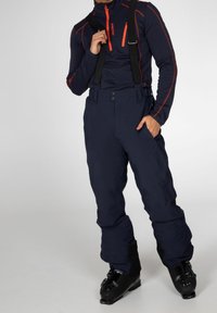 Personne portant un pantalon de ski bleu marine avec des bretelles noires, une veste zippée bleu marine et orange et des chaussures de ski noires, debout.