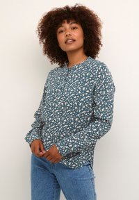 Chemise à manches longues en teal avec un motif abstrait blanc, encolure ronde et coupe décontractée. Assortie à un jean en denim bleu. Tissu texturé.