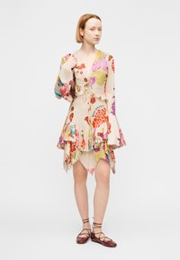 Φόρεμα με λουλουδάτο μοτίβο σε ανοιχτό μπεζ ύφασμα, με μακριά φαρδιά μανίκια, δέσιμο μπροστά και στρώματα στο τελείωμα με πολύχρωμες λεπτομέρειες.