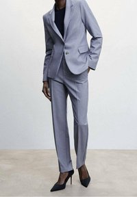 Costume ajusté gris clair avec une veste à un bouton et un pantalon à jambes droites. Tissu texturé, associé à des escarpins noirs à talons pointus.