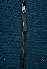 Mörkblå jacka i strukturerad tyg, med en central dragkedja, tryckknappar nedtill och en märkesdragkedjehängare.