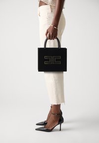 Sac en cuir noir avec logo doré, de forme rectangulaire petite et avec des poignées supérieures. Porté avec un pantalon blanc court et des escarpins noirs.