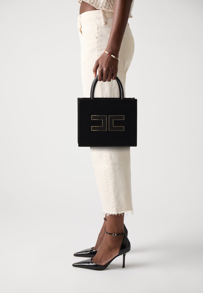 Sac en cuir noir avec logo doré, de forme rectangulaire petite et avec des poignées supérieures. Porté avec un pantalon blanc court et des escarpins noirs.