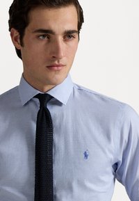 Camicia a scacchi blu chiaro con colletto a punta, caratterizzata da un piccolo logo ricamato blu sul petto e una cravatta nera a trama.