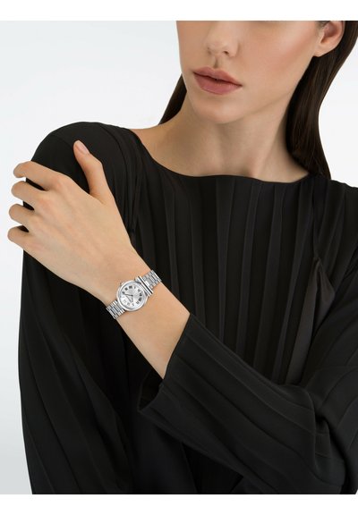 Silver wristwatch with a round face, Roman numerals, and a textured band, worn on a hand with a pleated black sleeve visible.