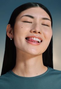 Gros plan d'une femme aux longs cheveux noirs, portant un haut teal, souriant les yeux fermés, mettant en valeur des lèvres brillantes et des boucles d'oreilles en argent.