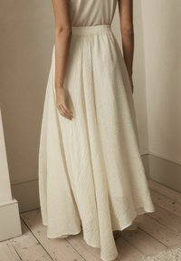 Longue jupe crème fluide avec une surface texturée et une taille élastique. La jupe tombe gracieusement jusqu'au sol, avec des plis subtils.