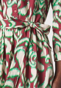 Gros plan d'une ceinture nouée à la taille sur une robe à manches longues avec un motif abstrait en spirale verte et rouge sur tissu crème.