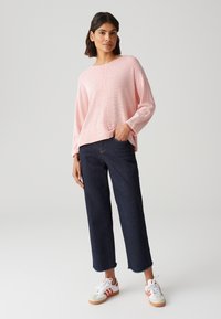 Rosa strukturiertes Strickpullover mit rundem Ausschnitt, kombiniert mit dunkelblauen, geraden Jeans und weißen Sneakers mit roten Akzenten.
