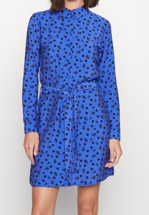 Kobieta ubrana w niebieską sukienkę koszulową z długimi rękawami, czarnym kwiatowym wzorem i przewiązanym paskiem w talii, stojąca na jednolitym tle.