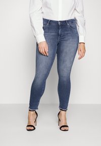 Jeans en denim bleu skinny avec une finition délavée, dotés de poches avant et d'un ourlet avec un subtil détail effiloché. Associés à des sandales à talons hauts noires.