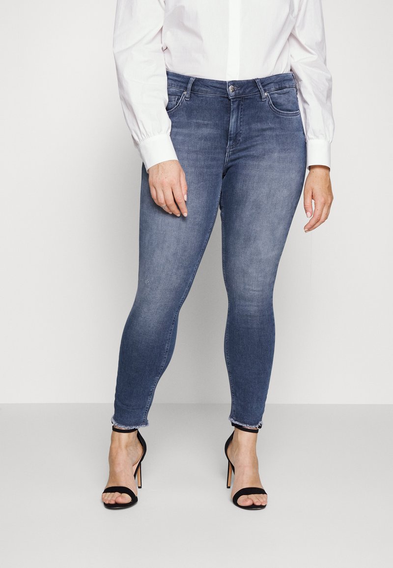 Jeans en denim bleu skinny avec une finition délavée, dotés de poches avant et d'un ourlet avec un subtil détail effiloché. Associés à des sandales à talons hauts noires.