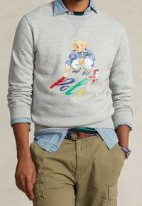 Grå huvtröja med färggrafik av en björn, med ordet "Polo". Lager över en ljusblå denimskjorta, tillsammans med olivgröna cargobyxor.