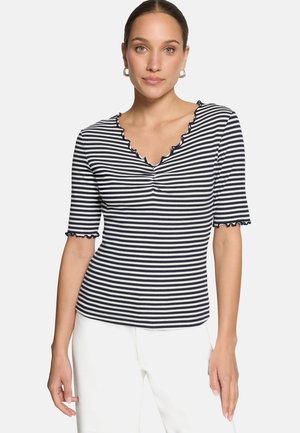 Femme portant un haut à rayures horizontales noires et blanches avec un décolleté en V à volants et des manches courtes, associée à un pantalon blanc, faisant face à l'avant.