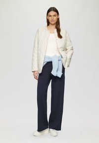 Gequilte ivoorkleurige bomberjack over een lichte top, gecombineerd met marineblauwe wijde broek en een lichtblauwe gebreide trui om de taille; witte platform sneakers.