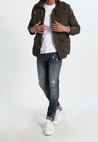 Olivengrøn militærinspireret jakke med flere lommer, hvid t-shirt, slidte blå jeans og hvide sneakers. Stående stilling.