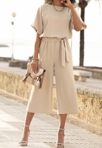 Mono beige con piernas anchas, mangas cortas y un cinturón a juego. Combinado con un bolso texturizado y plegable y tacones color arena con tiras.