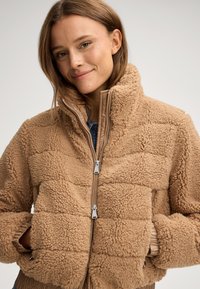 Beżowa polarowa kurtka z teksturowaną, pikowaną powierzchnią, zapinana na zamek, z wysokim kołnierzem i elastycznymi mankietami, posiadająca dwie boczne kieszenie.