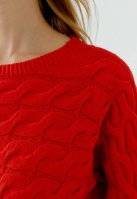 Roter Strickpullover mit strukturiertem Wellenmuster, geripptem Ausschnitt und weichem Stoff. Nahaufnahme hebt Design- und Farbdetails hervor.