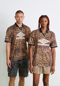 Du žmonės, apsirengę atitinkančiais leopardų rašto Umbro polo marškiniais: vienas su juodais šortais, kitas su smėlio spalvos sijonu, stovi prieš baltą foną.