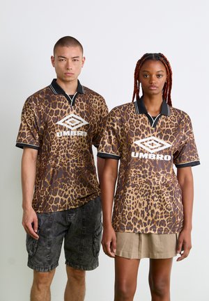 Two people wearing matching leopard print Umbro polo shirts, one paired with black shorts, the other with a beige skirt, standing against white.
