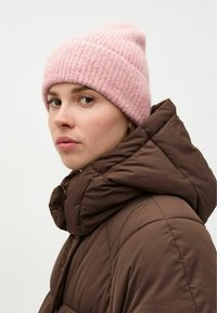 Světle růžová pletená čepice s jemným vzorem, nošená s hnědou péřovou bundou s vysokým límcem a prošívaným designem.