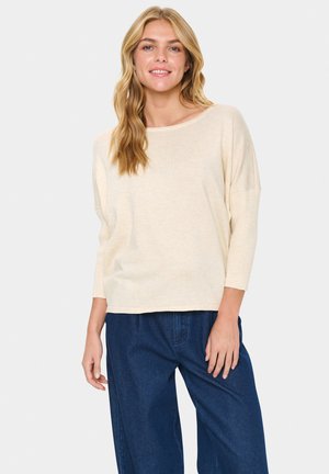 Haut beige à manches longues avec un large décolleté et une texture douce, associé à un jean bleu foncé ample. Design décontracté et confortable.