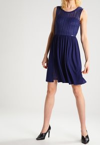 Marineblaues Kleid mit strukturiertem Oberteil und fließendem Rock. Ärmelloses Design, knielang und kombiniert mit schwarzen High Heels.