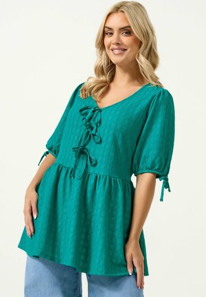 Kvinde med blondt hår iført en teal tekstureret bluse med tre bindebånd foran og puffede ærmer, kombineret med lyseblå jeans, smilende blidt.